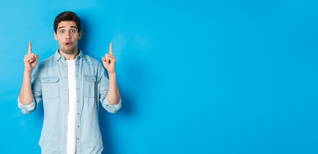 Photo gratuite portrait d'un bel homme étonné pointant les doigts vers le haut montrant le logo sur fond bleu