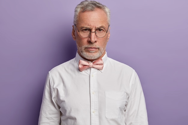 Portrait De Bel Homme Européen Aux Cheveux Gris Regarde Avec Une Expression De Visage Stricte à Travers Des Lunettes Rondes, Porte Une Chemise Blanche Formelle Et Noeud Papillon