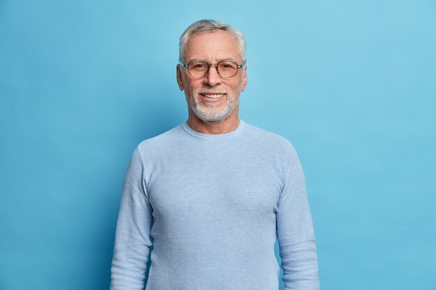 Portrait de bel homme européen barbu aux cheveux gris et à la barbe sourit agréablement regarde directement à l'avant étant de bonne humeur a de la chance porte des lunettes et un pull isolé sur un mur bleu