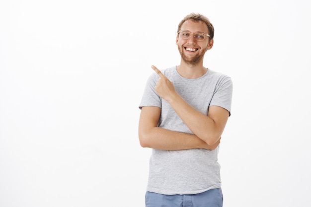 Portrait d'un bel homme européen insouciant optimiste avec des soies dans des verres souriant et riant avec désinvolture ayant une bonne journée pointant vers le coin supérieur gauche avec un regard satisfait et ravi sur un mur blanc