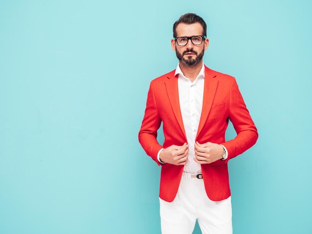 Portrait d'un bel homme hipster élégant confiant modèle sexuel sexy moderne vêtu d'un élégant costume rouge Mode homme posant en studio près du mur bleu à lunettes