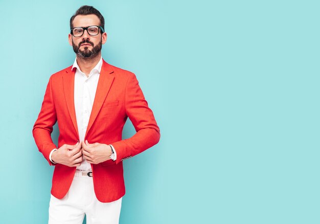 Portrait d'un bel homme hipster élégant confiant modèle sexuel sexy moderne vêtu d'un élégant costume rouge Mode homme posant en studio près du mur bleu à lunettes