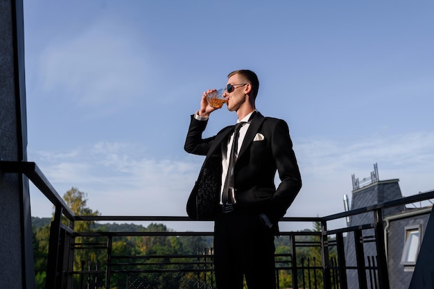 Photo gratuite portrait d'un bel homme marié en costume noir élégant debout sur le balcon en train de boire en regardant le coucher du soleil célébrer le jour du mariage stag party