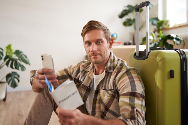 Photo gratuite portrait d'un bel homme souriant avec une valise qui regarde un smartphone et tient des billets de voyage et un passeport