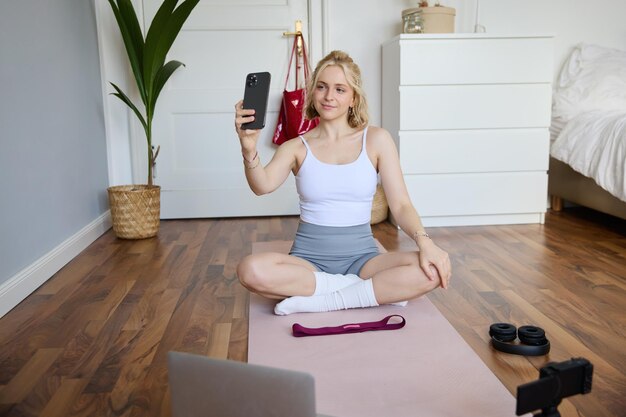 Photo gratuite portrait d'une belle blogueuse de style de vie qui s'enregistre en train de s'entraîner à la maison assise sur un tapis en caoutchouc