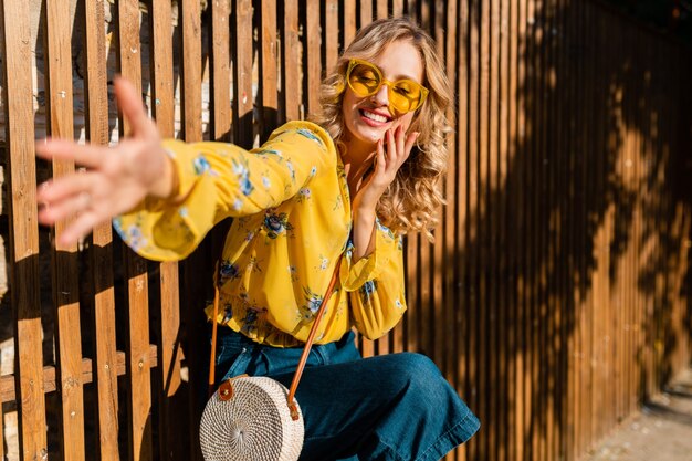 Portrait de belle blonde émotionnelle en riant élégante femme souriante en chemisier jaune portant des lunettes de soleil, sac à main en paille de style bali