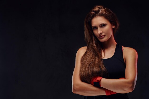 Portrait d'une belle boxeuse sportive en vêtements de sport et mains bandées, posant avec les bras croisés. Isolé sur un fond texturé sombre.