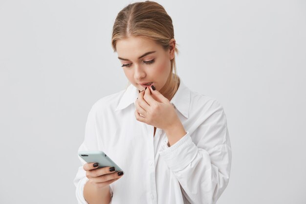 Portrait de la belle étudiante caucasienne habillée avec désinvolture tenant un téléphone portable, communiquant avec des amis via les réseaux sociaux, utilisant une connexion Internet élevée, touchant ses lèvres.