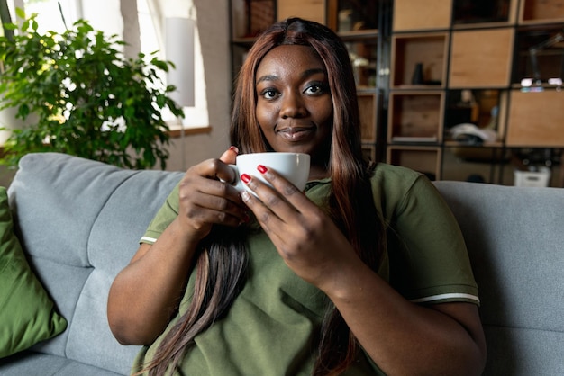 Portrait d'une belle femme africaine assise sur un canapé et buvant du thé isolé sur l'intérieur de la maison