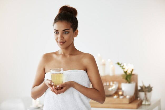 Portrait de la belle femme africaine en serviette tenant le verre avec du thé en souriant.