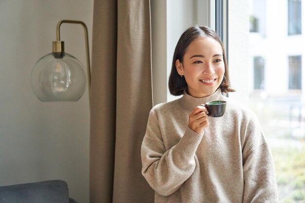 Portrait d'une belle femme asiatique assise à la maison avec une tasse de café en dégustant son expresso tout en regardant par la fenêtre extérieure un passant souriant joyeusement