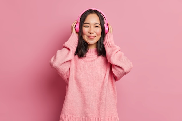 Portrait de la belle femme asiatique meloman porte des casques stéréo électroniques sans fil écoute la piste audio préférée ou la chanson populaire recrée avec de la bonne musique aime la mélodie tranquille porte un pull rose