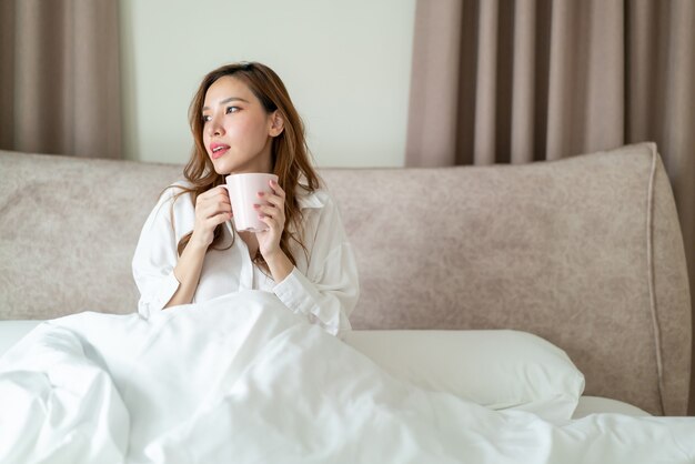 Portrait belle femme asiatique se réveiller et tenant une tasse de café ou une tasse sur le lit le matin