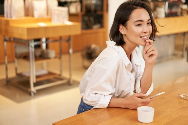 Portrait d'une belle femme asiatique avec un smartphone se relaxant dans un café assis et savourant un café tout en