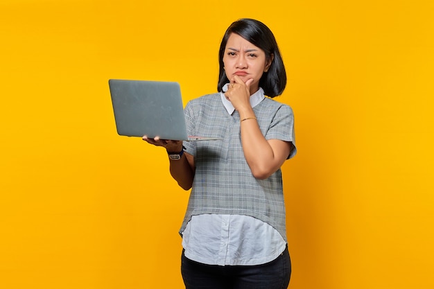 Portrait d'une belle femme asiatique tenant un ordinateur portable et pensant à quelque chose avec la main sur le menton et regardant la caméra sur fond jaune