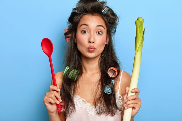 Portrait de belle femme au foyer garde les lèvres arrondies, veut embrasser son mari, tient une cuillère et des légumes frais verts