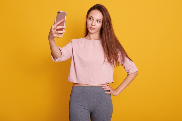 Portrait de belle femme aux cheveux bruns avec des cheveux raides en pantalon gris et chemise rose prenant selfie