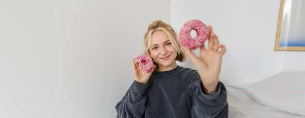 Photo gratuite portrait d'une belle femme blonde montrant deux beignets roses délicieuses pâtisseries mangeant le dessert et