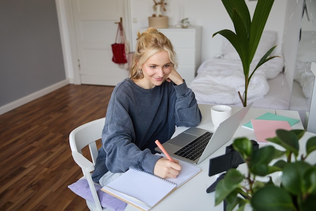 Photo gratuite portrait d'une belle femme blonde souriante qui écrit des notes, fait ses devoirs, étudie à domicile.