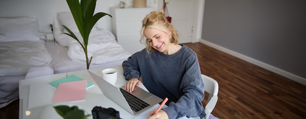 Photo gratuite portrait d'une belle femme blonde souriante qui écrit des notes, fait ses devoirs, étudie à domicile.