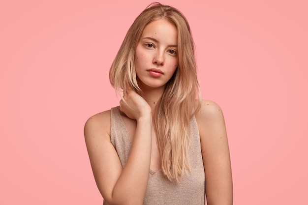 Photo gratuite portrait de belle femme blonde touche le cou avec les mains, regarde mystérieusement et sérieusement, a la peau pure, va avoir une promenade en plein air, des modèles contre le mur rose