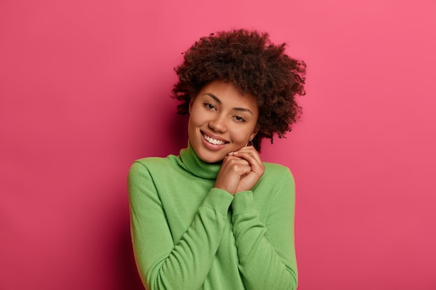 Portrait de belle femme bouclée incline la tête, se sent heureuse de passer le week-end avec sa famille, garde les mains pressées ensemble, porte un cavalier vert