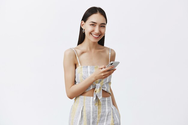 Portrait de belle femme bronzée sympathique et charmante en haut assorti avec un short, tenant un smartphone et regardant joyeusement, aimant le nouveau téléphone sur un mur gris