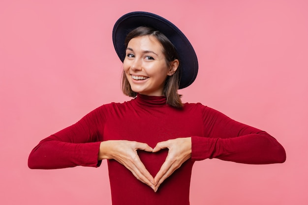 Portrait de la belle femme brune au chapeau, montre le geste du cœur sur la poitrine, être passionné, exprimer son amour à la personne proche, se dresse.