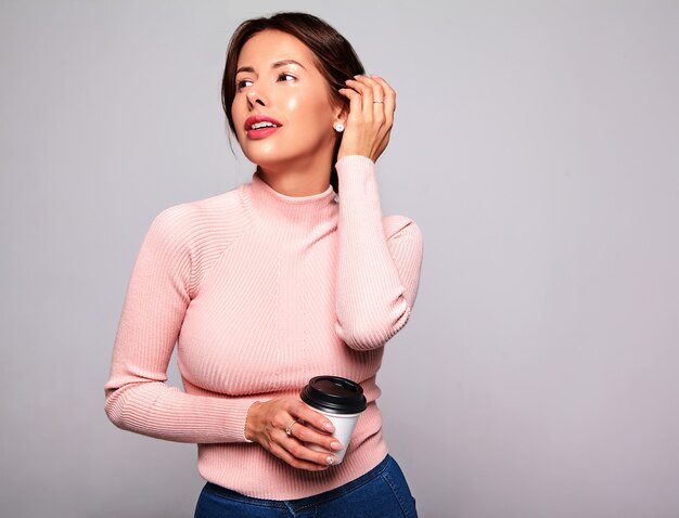 Portrait de la belle femme brune mignonne modèle dans des vêtements décontractés d'été rose sans maquillage isolé sur gris. Buvant du café