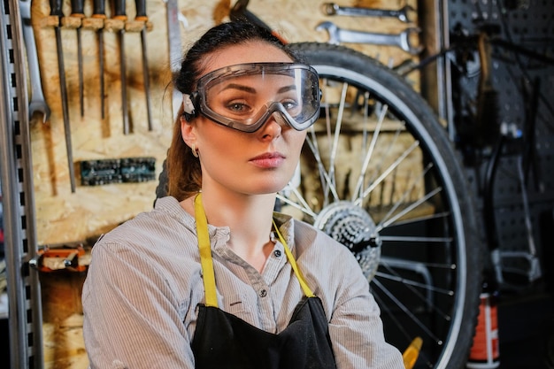 Portrait d'une belle femme brune portant des vêtements de travail, un tablier et des lunettes, debout dans un atelier contre des outils muraux.