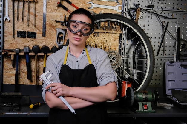 Photo gratuite portrait d'une belle femme brune portant des vêtements de travail, un tablier et des lunettes, tient un étrier, debout dans un atelier contre des outils muraux.