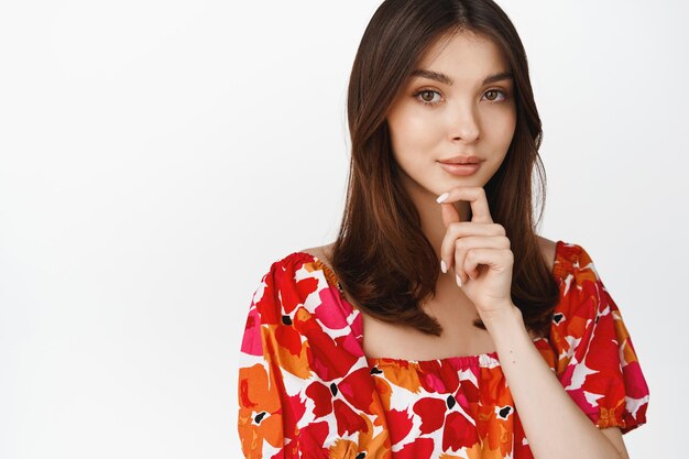 Portrait d'une belle femme brune regardant la caméra pensive portant une élégante robe à fleurs debout sur fond blanc