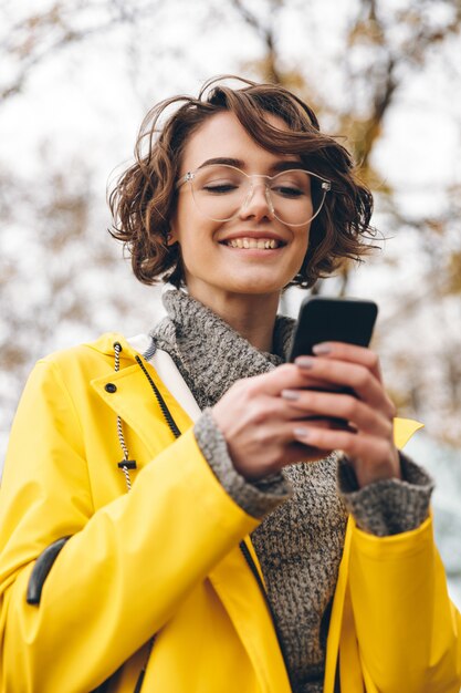 Portrait de la belle femme brune taper un SMS ou faire défiler le flux dans le réseau social à l'aide de son smartphone tout en étant en plein air