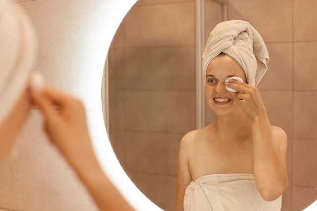 Portrait d'une belle femme caucasienne en serviette sur la tête regardant dans un miroir et nettoyant son visage avec un coton, enlevant le maquillage de ses yeux, souriant et exprimant le positif.