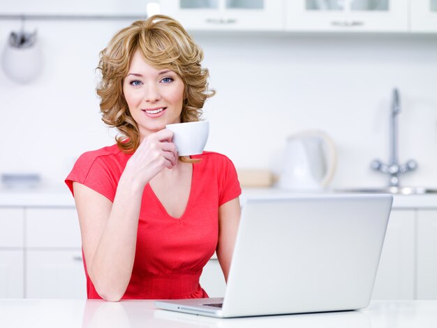 Portrait de la belle femme décontractée assis dans la cuisine avec une tasse de café et un ordinateur portable