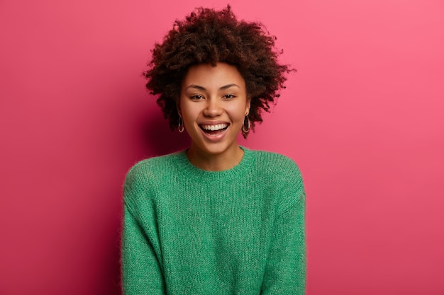 Portrait de belle femme ethnique bouclée sourit largement, profite d'une journée de congé, a une conversation heureuse avec son interlocuteur, discute de la préparation des vacances, porte un pull vert, isolé sur un mur rose.