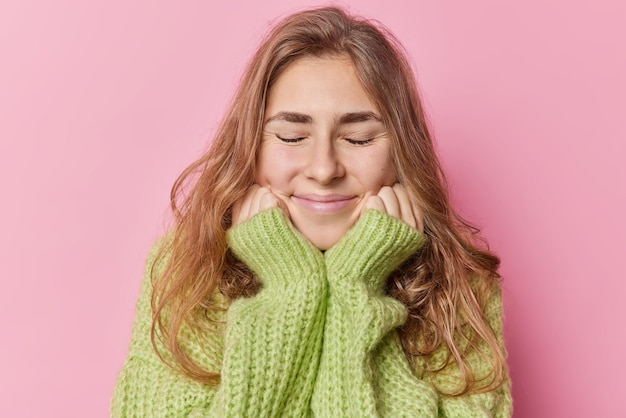 Portrait d'une belle femme européenne aux cheveux longs garde les mains sous le menton a une expression faciale douce ferme les yeux de la satisfaction rêverie porte un pull tricoté chaud isolé sur fond rose.