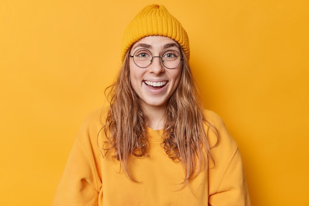 Portrait d'une belle femme européenne heureuse aux cheveux longs yeux bleus blancs même dents porte des lunettes rondes transparentes chapeau et sweat-shirt isolés sur fond jaune Émotions positives
