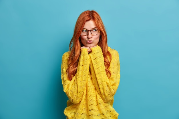 Portrait de belle femme européenne rousse garde les mains sous le menton et les lèvres arrondies rêve de quelque chose de romantique porte des lunettes transparentes et un pull jaune.