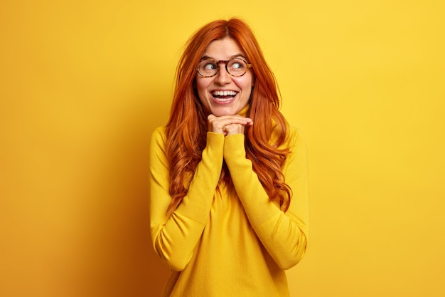 Portrait de belle femme européenne rousse sourit largement garde les mains sous le menton regarde de côté porte joyeusement cavalier occasionnel exprime le bonheur.