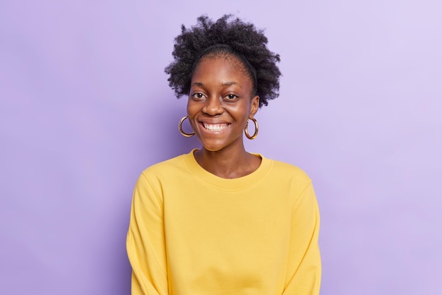 Portrait d'une belle femme gaie à la peau sombre avec des cheveux bouclés sourit à pleines dents portant un pull jaune décontracté a une bonne humeur isolée sur fond violet. Concept d'émotions et de sentiments heureux