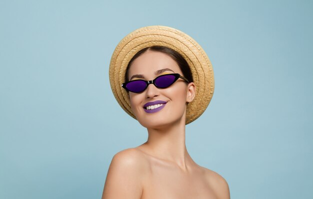 Portrait de belle femme avec maquillage lumineux, chapeau et lunettes de soleil sur fond bleu studio. Marque et coiffure élégantes et à la mode. Couleurs de l'été. Beauté, mode, concept publicitaire. Regarde de côté.