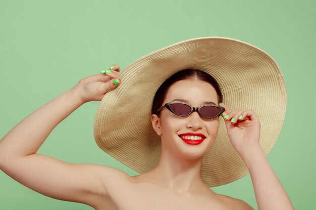 Portrait De Belle Femme Avec Maquillage Lumineux, Chapeau Et Lunettes De Soleil Sur Fond De Studio Vert. Marque Et Coiffure élégantes Et à La Mode. Couleurs De L'été. Concept De Beauté, De Mode Et De Publicité. Souriant.