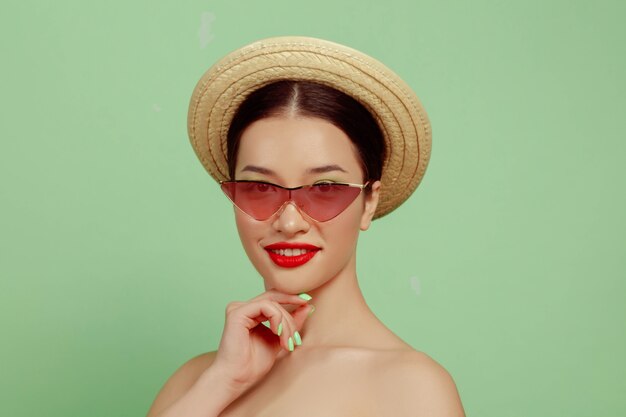 Portrait de belle femme avec maquillage lumineux, lunettes rouges et chapeau sur fond vert studio. Marque et coiffure élégantes et à la mode. Couleurs de l'été. Concept de beauté, de mode et de publicité. Posant.