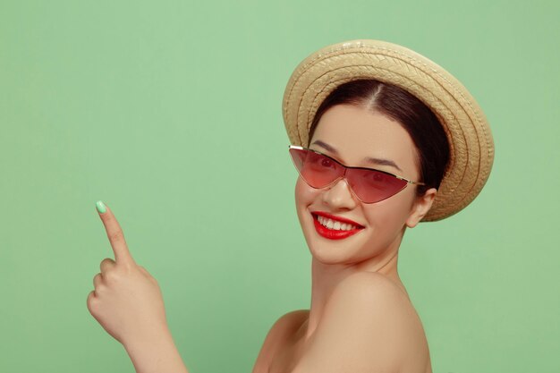 Portrait de belle femme avec maquillage lumineux, lunettes rouges et chapeau. Marque et coiffure élégantes et à la mode. Couleurs de l'été. Montrer du doigt.