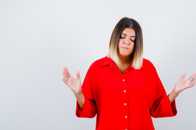 Portrait d'une belle femme montrant un geste impuissant, regardant vers le bas en blouse rouge et regardant la vue de face contrariée