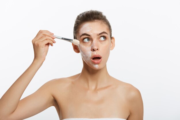 Portrait belle femme nue appliquer un masque cosmétique à l'argile blanche avec un pinceau isolé sur un dos blanc