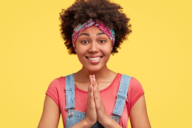 Portrait de la belle femme à la peau foncée avec des cheveux croquants, a une expression implorante, garde les mains dans un geste de prière, porte une salopette en denim sourit largement, isolée sur un mur jaune. Le langage du corps