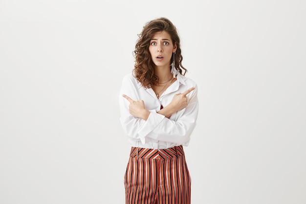 Portrait d'une belle femme réussie aux cheveux bouclés dans des vêtements à la mode pointant dans les deux sens et étant perplexe ou dévasté debout contre un mur gris