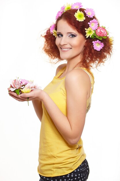 Portrait de la belle femme rousse au gingembre souriant en tissu jaune avec des fleurs colorées rose jaune dans les cheveux isolé sur blanc tenant des fleurs